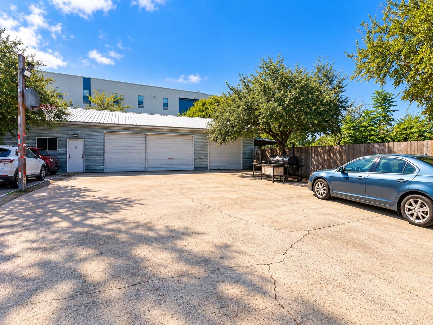 Outdoor lot with a smoker and BBQ pit under shade trees beside the studio building and parked cars